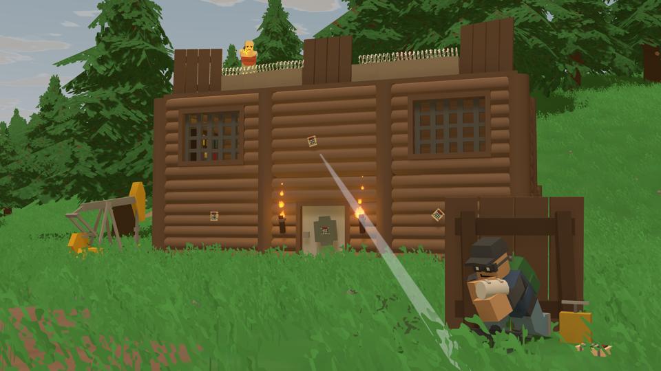 A player behind cover applies a bandage while another player standing on a house shoots near them. The house is covered in explosives, ready for the healing player to detonate them.